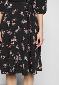 Svart klänning med blommönster som visar rosa blommor och känsliga fåglar. Inkluderar knytbälte i midjan och en flödande kjol med strukturerat tyg.