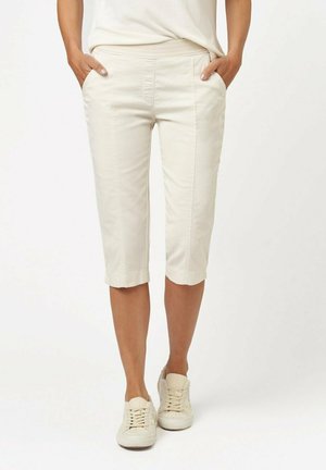 Pantaloni cropped beige chiaro con una texture liscia, tasche frontali e un dettaglio di cucitura sottile lungo le gambe, abbinati a sneakers bianche.