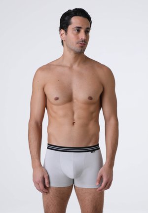 Uomo adulto in forma con capelli corti e scuri che indossa boxer grigio chiaro con una fascia nera in vita, in piedi davanti a uno sfondo chiaro e semplice.