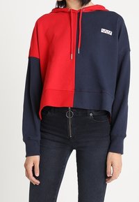 Mujer que lleva una sudadera corta a rayas rojas y azul marino con un parche que dice "AMOR", combinada con unos jeans negros de tiro alto con un tirador de cremallera circular.