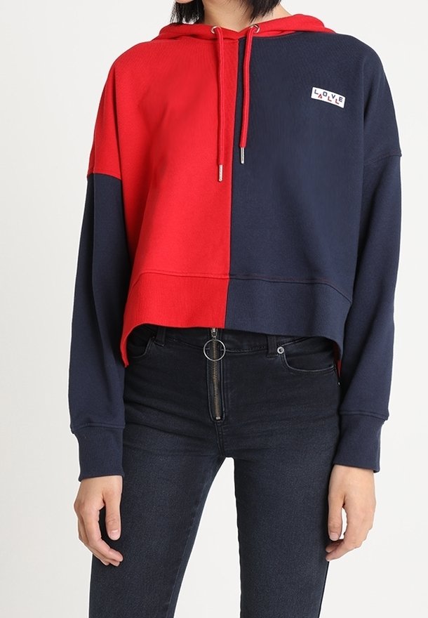 Mujer que lleva una sudadera corta a rayas rojas y azul marino con un parche que dice "AMOR", combinada con unos jeans negros de tiro alto con un tirador de cremallera circular.