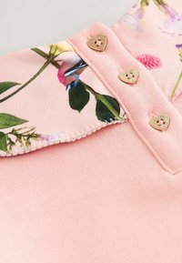Camisa polo de suave tejido rosa con un cuello estampado con flores. Presenta tres botones dorados en forma de corazón para el cierre. Textura suave.