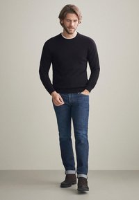 Svart stickad tröja med rund halsringning, i kombination med blå denimjeans. Personen bär bruna ankelstövlar och har uppvikta ärmar på jeansen.