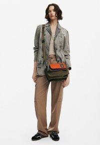Grön tygaxelväska med orange och lila accenter. Har dragkedjestängning och logodetalj. Bärs med beige cargo-byxor och en denimjacka.