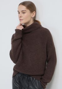 Donna con capelli castano chiaro che indossa un maglione a collo alto marrone a costine e pantaloni in pelle nera, che posa in modo pensieroso contro un muro bianco.