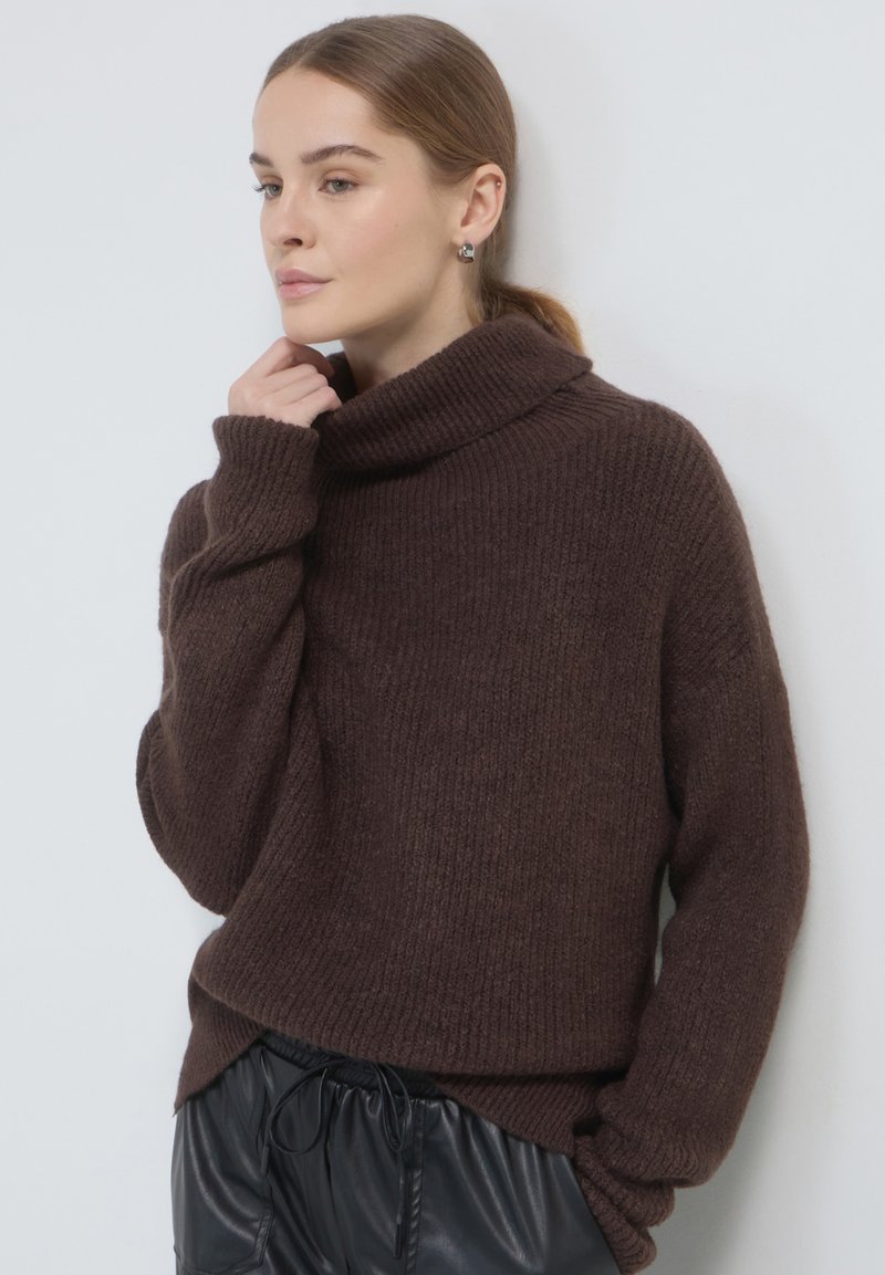 Donna con capelli castano chiaro che indossa un maglione a collo alto marrone a costine e pantaloni in pelle nera, che posa in modo pensieroso contro un muro bianco.