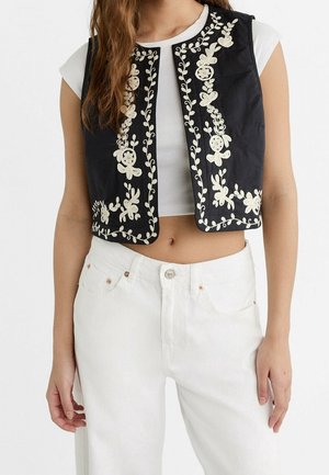 Woman wearing a black sleeveless vest with white floral embroidery over a white cropped top and white high-waisted pants.