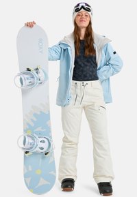 Snowboard mit weißer Oberseite, blauen Akzenten und Wellenmustern. Modell trägt eine blaue Jacke, ein schwarzes gemustertes Hemd und cremefarbene Hosen mit Reißverschlüssen.