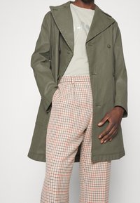 Muž v olivově zeleném trenčkotě, světle béžovém tričku s potiskem a béžových kostkovaných kalhotách s červeným a černým čtvercovým vzorem.