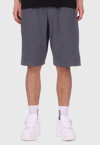 Graue, gerippte Shorts mit einem elastischen Bund, ausgestattet mit seitlichen Taschen und einem kleinen, gestickten Logo am linken Bein. Getragen mit weißen Turnschuhen.