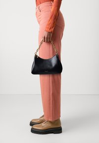 Black leather handbag with gold-tone chain handle, curved shape, and gold foil logo. Paired with pink pants and brown lace-up shoes.