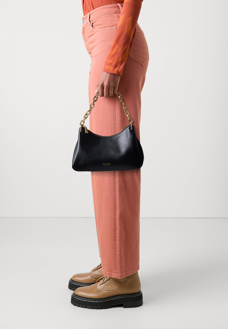 Black leather handbag with gold-tone chain handle, curved shape, and gold foil logo. Paired with pink pants and brown lace-up shoes.