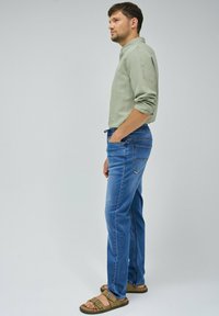 Camisa de manga larga verde claro, jeans azules con detalles de costura y sandalias marrones. El atuendo tiene un corte holgado y un aspecto casual.