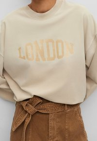 Beige collegepaita, jossa "LONDON" hieman tummemmalla sävyllä, yhdistettynä ruskeaan, teksturoituun korkeavyötäröiseen vyöhön. Yksinkertainen, rento istuvuus.