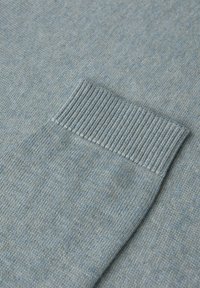 Ljusblå stickad tröja med ribbad manschett. Tyget uppvisar en subtil melerad textur som skapar djup och variation i färgen.