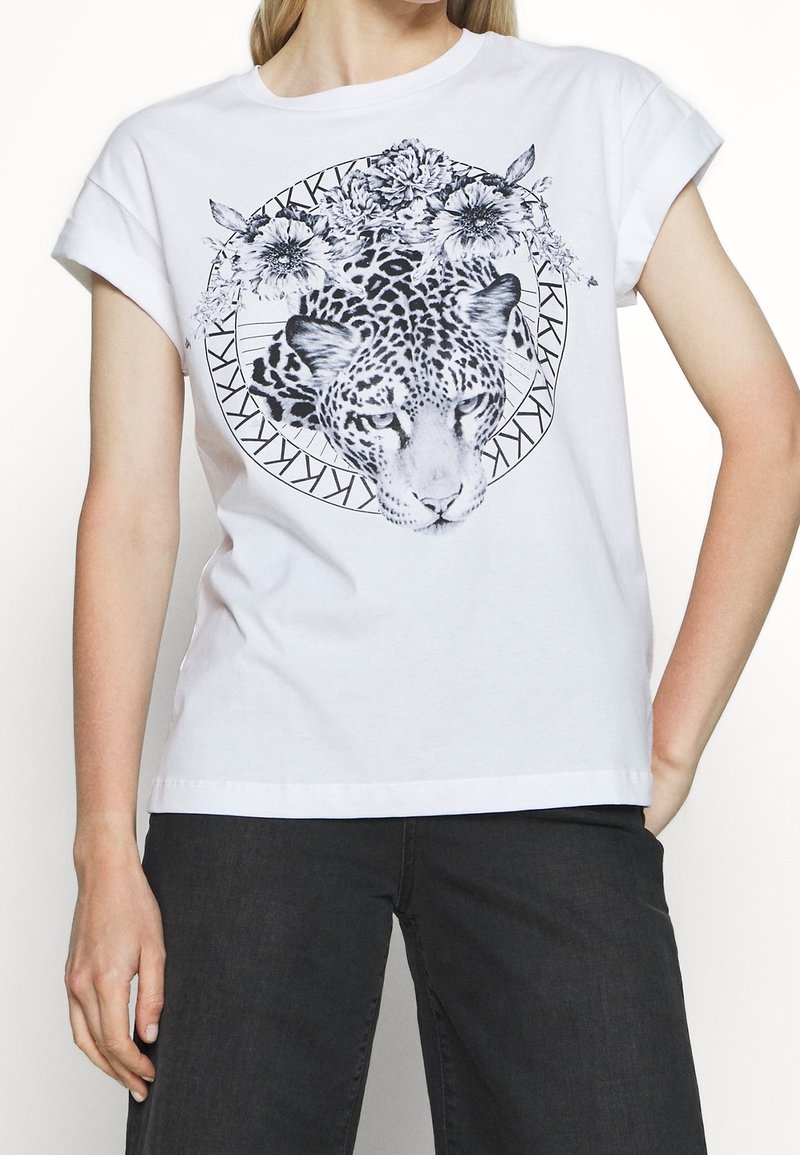 Camiseta de algodón blanca con un diseño de estampado de leopardo negro enmarcado por patrones florales y geométricos, con mangas enrolladas y un corte relajado.