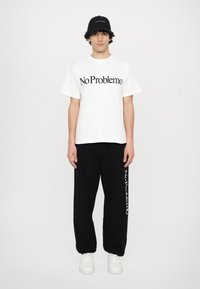 White t-shirt with black "No Problemo" print, paired with loose black sweatpants featuring a similar print. Worn with white sneakers. Black cap.