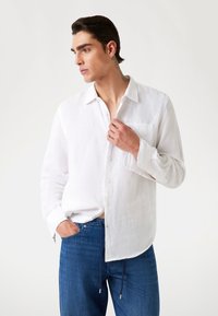 Camisa de lino blanca de manga larga con botones en la parte delantera, un bolsillo en el pecho, textura suave y bajo curvado, combinada con pantalones de mezclilla azules con cordón.