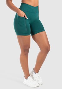 Szorty w kolorze turkusowym, o wysokim stanie, wykonane z elastycznego materiału, z bocznymi kieszeniami i akcentami w postaci szwów. Noszone z białymi butami sportowymi.