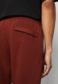Burgundy sweatpants med en elastisk midja, som har en liten bakficka och mjukt, strukturerat tyg, delvis visade med en hand.