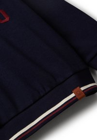 Detailní záběr na spodní lem tmavě modrého sweatshirtu s bílo-červenými pruhovanými náplety a malou hnědou koženou nášivkou s vyraženým logem.