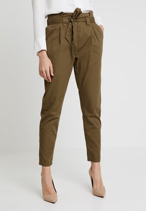 Kvinde iført olivengrønne, højtaljede paperbag-bukser med bindebælte, hvid bluse med lange ærmer og beige spidse hæle, stående mod en ensfarvet baggrund.