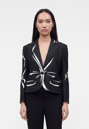Woman with long black hair wearing black blazer with white abstract line designs, standing against plain light background.