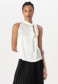 Blouse sans manches en soie blanche avec un col haut et un détail à nouer. Le modèle la porte avec une jupe plissée noire, mettant en valeur la texture lisse et le design élégant.