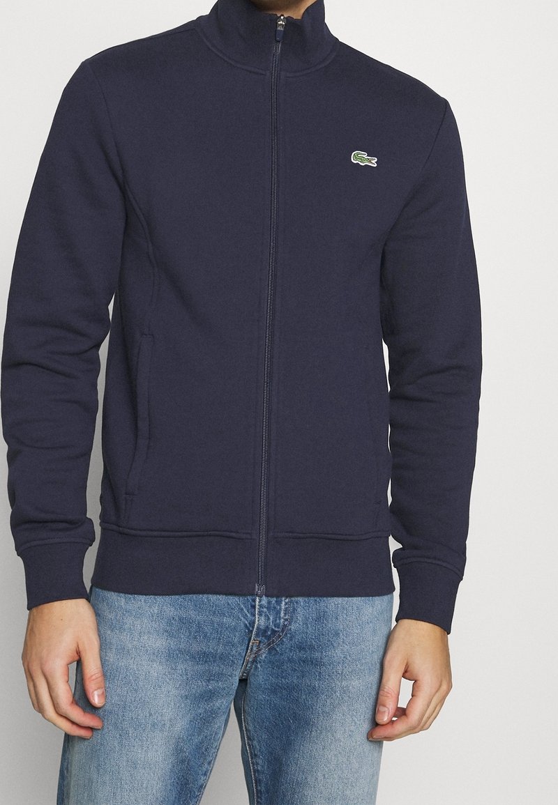 Uomo che indossa una giacca zip-up blu navy con un piccolo logo di coccodrillo verde sul petto e jeans di denim blu chiaro.