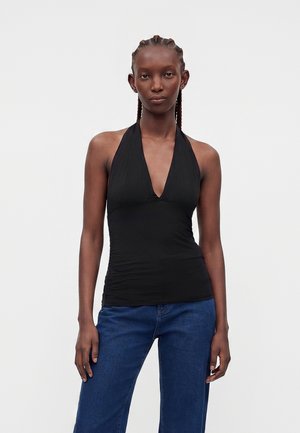 Femme aux cheveux tressés portant un haut noir à col halter et un jean bleu, debout devant un fond uni gris clair.