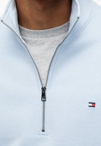 Personne portant une veste zippée bleu clair par-dessus un t-shirt gris, avec la fermeture éclair partiellement ouverte et un petit logo sur la poitrine de la veste.