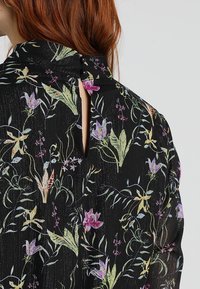 Schwarze Bluse mit Blumenmuster aus leichtem Stoff, mit mehrfarbigen Blumenmustern, einem Schlüsselloch-Rücken und Knopfverschlüssen am Hals.