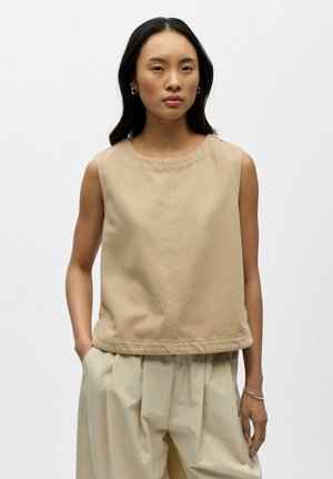 Femme aux longs cheveux foncés portant un haut beige sans manches et un pantalon ample beige clair, debout avec une main dans la poche, fond neutre.