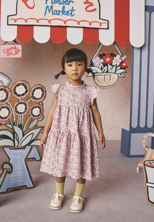 Ung pige med fletninger iført en blomsterkjole og gule sokker står foran et baggrundstæppe af papblomster til et marked.