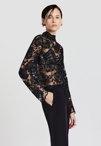 Schwarze transparente Bluse mit Blumenstickerei, langen Ärmeln, hohem Ausschnitt und zarten gewellten Manschetten, kombiniert mit schwarzen maßgeschneiderten Hosen.
