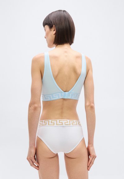 Γυναίκα, φαινομενικά από πίσω, φορώντας ανοιχτό γαλάζιο αθλητικό σουτιέν και λευκά εσώρουχα με χρυσές και μπλε γεωμετρικές λεπτομέρειες στη μέση.