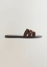 Sandale à glissière en daim marron avec deux larges sangles, présentant un accent en boucle dorée. Semelle plate avec une finition noire.