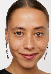 Närbild av en person med medelbrun hy, synliga fräknar och bruna ögon; iförd silverörhängen med en abstrakt design och en svart topp.