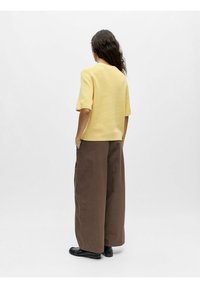 Haut jaune en tricot à manches courtes et pantalon marron ample, portés avec des chaussures plates noires ; vu de dos sur un fond clair.