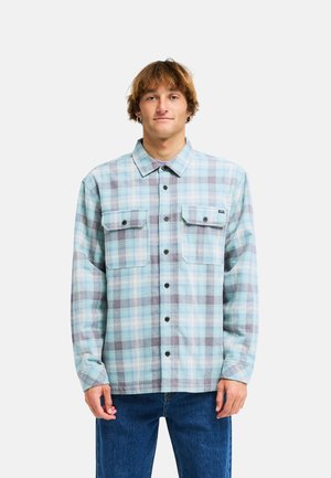 Giovane uomo con capelli castano chiaro che indossa una camicia a quadri azzurro chiaro e grigia abbottonata e jeans blu su uno sfondo semplice.