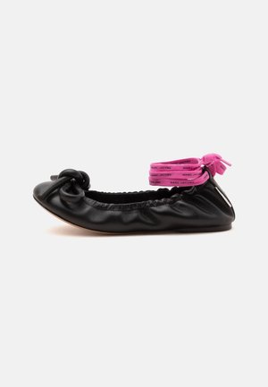 Ballerine plate en cuir noir avec des détails froncés, un nœud sur l’orteil et des brides de cheville roses avec du texte attachées autour du talon.