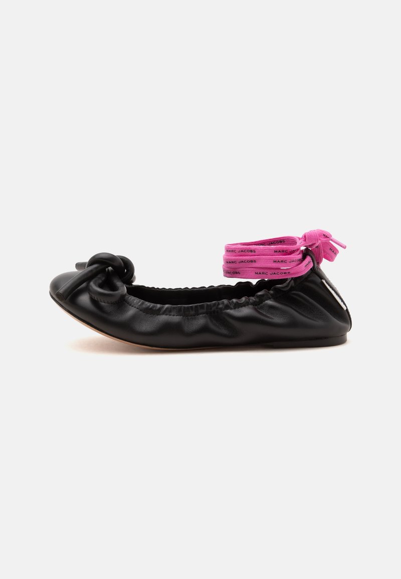 Ballerine plate en cuir noir avec des détails froncés, un nœud sur l’orteil et des brides de cheville roses avec du texte attachées autour du talon.
