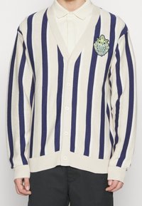 Knopf-Down-Strickjacke mit vertikalen blauen und cremefarbenen Streifen, featuring einen V-Ausschnitt, gerippte Ärmelbündchen und ein gesticktes Logo auf der linken Brust.