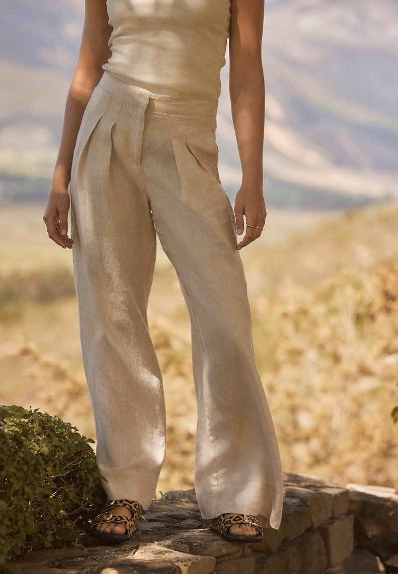 Femme portant un pantalon large en lin beige, un débardeur et des sandales imprimé léopard, debout dehors sur des marches en pierre.