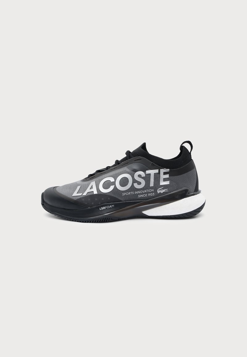 Μαύρο και γκρι αθλητικό παπούτσι Lacoste με λευκό κείμενο "LACOSTE" και λευκή μαξιλαρωμένη σόλα, με έλκηθρο στη φτέρνα και αθλητικά σχεδιαστικά στοιχεία.