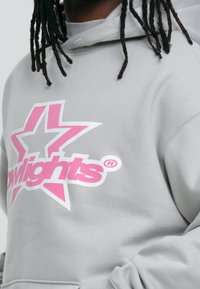 Sudadera con capucha gris de material suave que presenta un gran logo de estrella rosa y la palabra "Glowlights" en blanco. Incluye un bolsillo canguro.