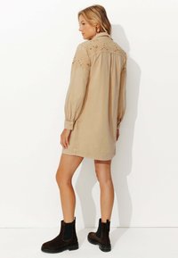 Robe chemise en denim beige avec broderies complexes sur les épaules, manches longues et coupe décontractée, associée à des bottines noires.