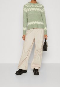 Zielony dzianinowy sweter w białą zygzakowatą wzór, zestawiony z luźnymi beżowymi spodniami cargo i czarnymi platformami. Mała czarna torebka w ręce.