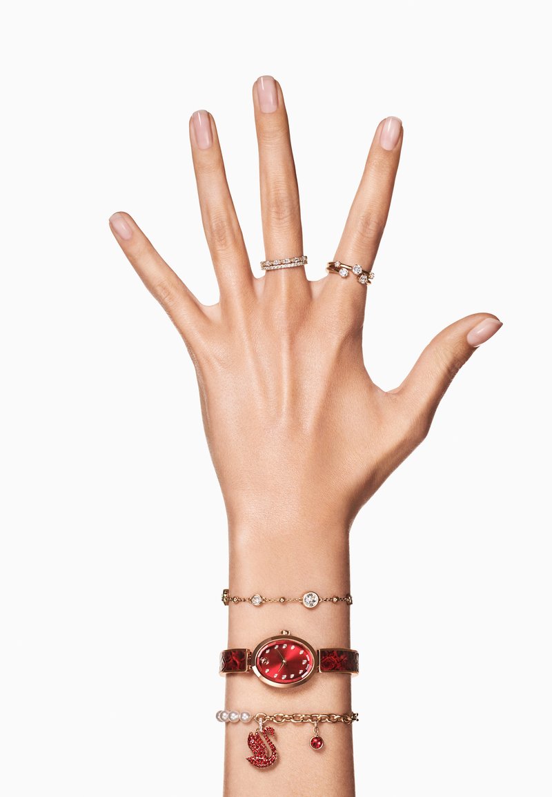 Relógio de pulso vermelho combinado com várias pulseiras, incluindo realces em prata e ouro. Anéis com pedras claras nos dedos, exibindo vários designs.