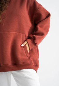 Weinroter Hoodie mit einer Fronttasche, gerippten Bündchen und Saum. Der Stoff wirkt weich mit einer glatten Textur, kombiniert mit weißen Hosen.